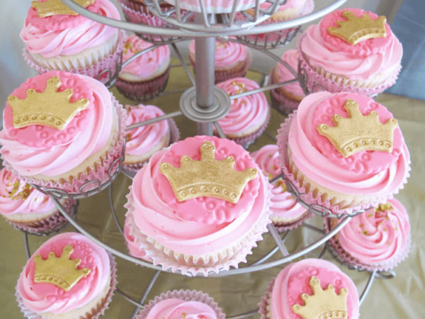 Pink cupcakes with pink frosting on a tiered cupcake stand. Each is topped with pink circles and gold crowns.